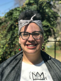 Alicia Bell, smiling in front of a tree and wearing tortoiseshell glasses, a gray headscarf, and a red earrings.