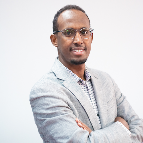 Headshot of Mukhtar Ibrahim smiling, wearing a white and gray checkered shirt, and gray suit jacket.