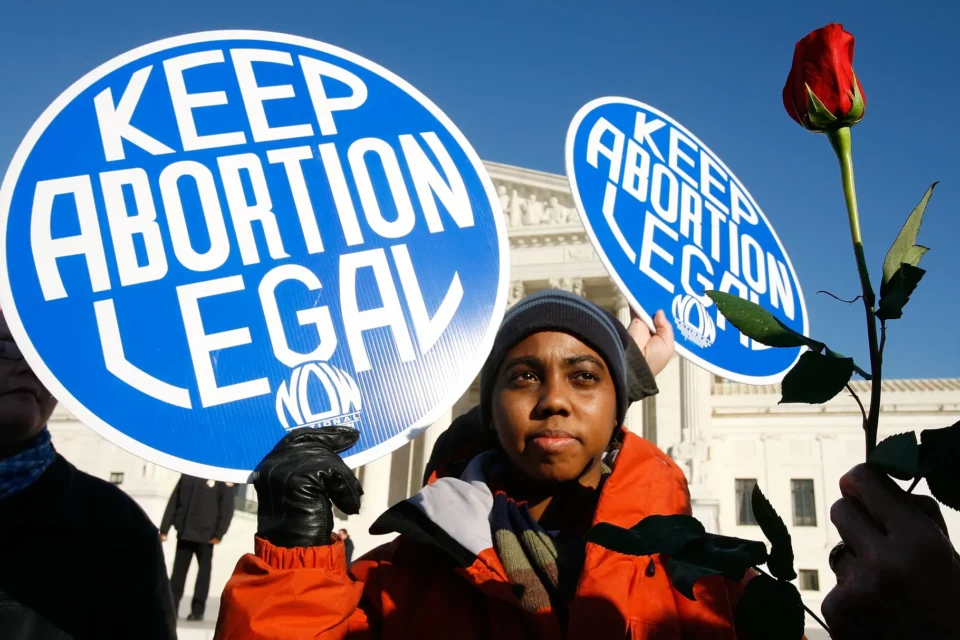 Organizer stands before the Supreme Court with a circular blue sign reading "KEEP ABORTION LEGAL." They are flanked by another sign and a red rose.