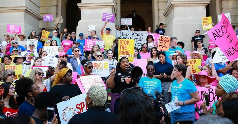 Dozens of diverse organizers stand in front of a government building while holding pink, yellow, and white pro-abortion and pro-reproductive justice signs. One organizer, at the center of the image, holds a megaphone.