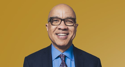 Ford Foundation President Darren Walker is pictured in a dar blue suit jacket, a light blue shirt, and a blue tie with red and white specks on it. He is wearing brown glasses and smiling, against a mustard yellow backdrop.