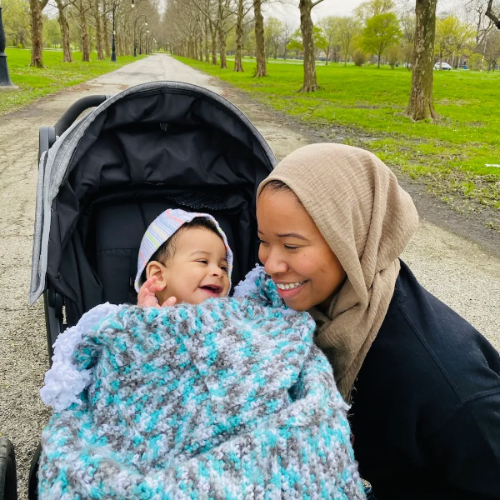 Makkah kneels down by her baby's carriage. Makkah wears a tan head scarf and black top. Her baby wears a stripped purple hoodie and is wrapped in a multi-colored blanket. Behind them is a long, tree-lined park path.