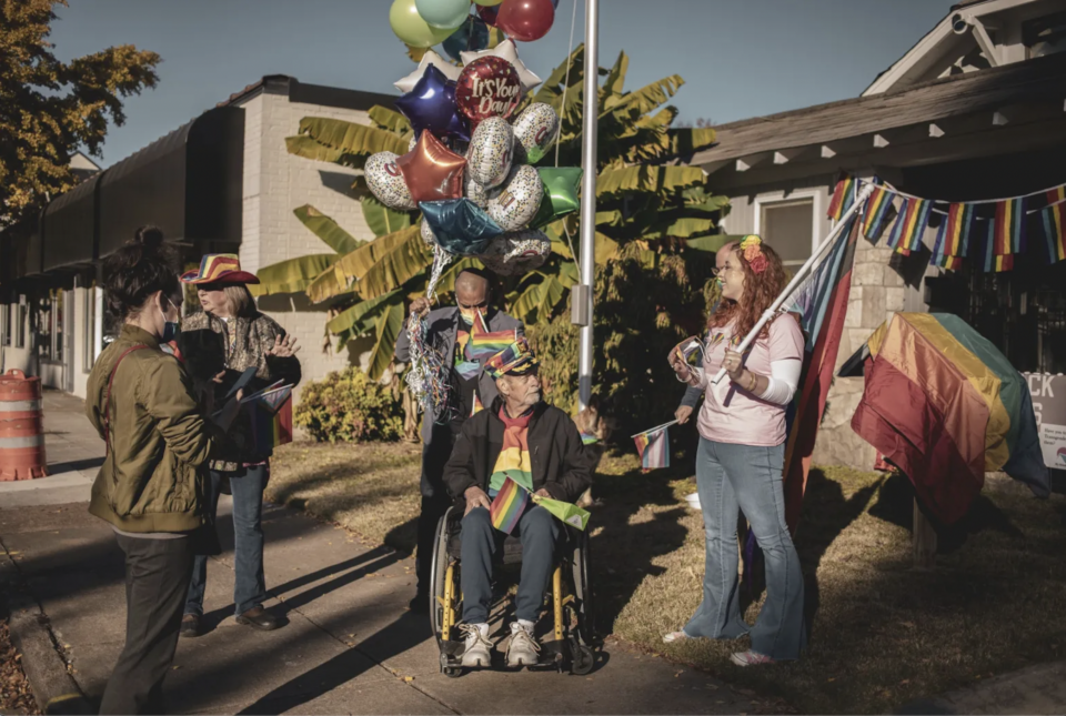 5 people surround 1 man sitting in a wheelchair on a building front lawn. They are all wearing pride gear. Around them are pride decorations and flags.