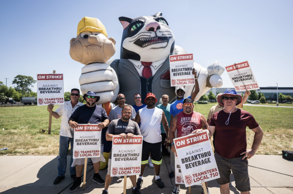 10 people stand in front of a balloon of an angry-looking cat in a business suit that holds a construction worker tightly in its left hand. The folks hold signs reading "On Strike against Breakthru Beverage. Teamsters Local 710." 