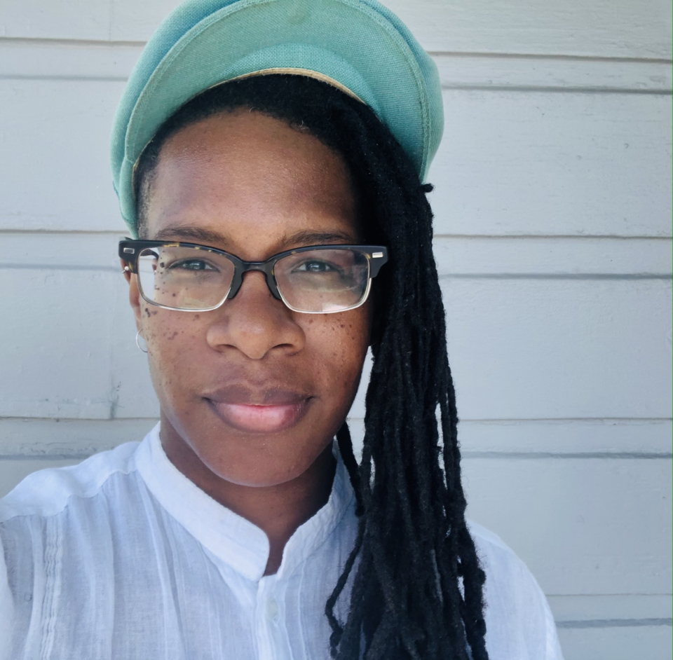 BLMF Senior Program Associate Jardyn Lake is pictured gazing into the camera. They have braids flowing down the right side of their face and shoulder. They're wearing a green cap, glasses, and white top. They stand in front of a white wall.
