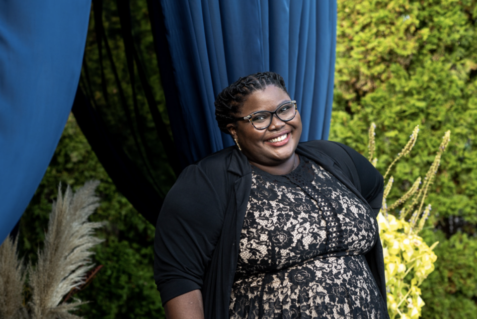 Bianca Carter smiling at the camera. She's wearing a black lace dress, glasses, and earrings. She's standing in front of two blue curtains and trees.