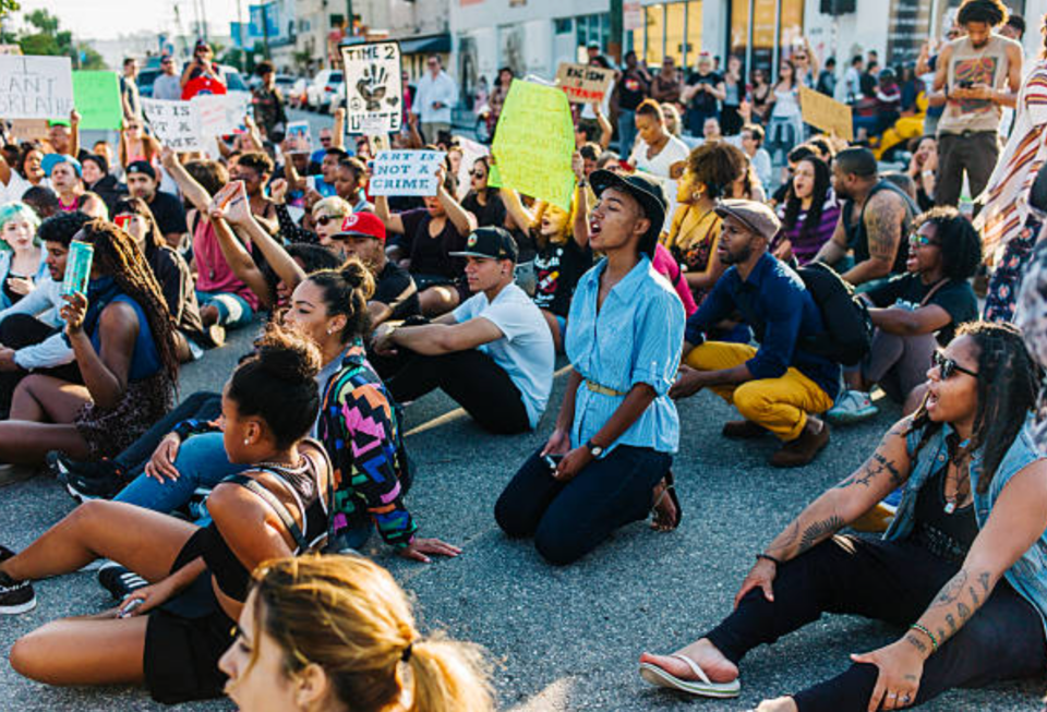 Folks sit, kneel, and hold up signs at a  rally action.