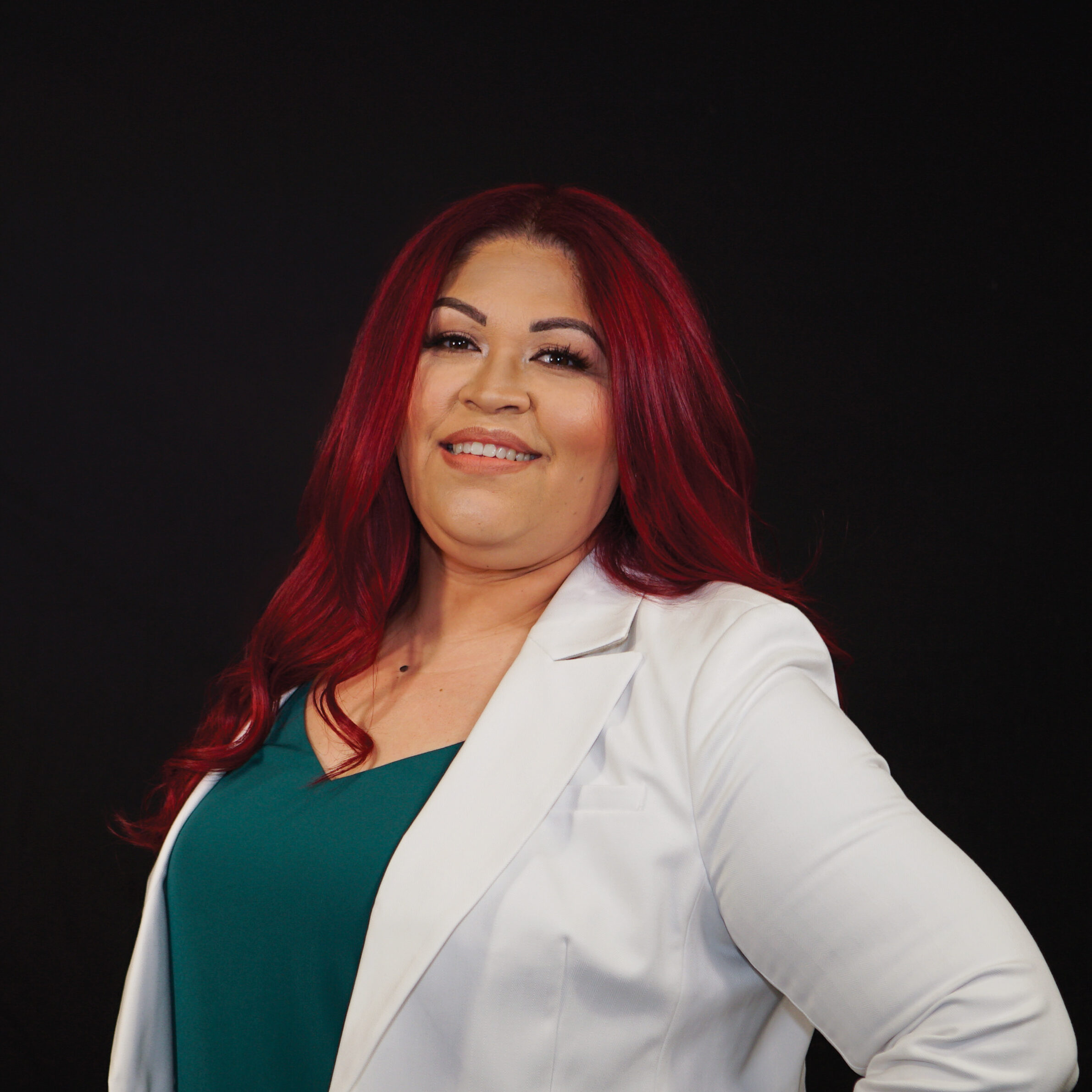 Headshot of Anna Hernandez against a black background. She has long, red hair and is wearing a green top under a white jacket. 