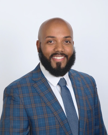 Headshot of Brandon Gleaton. He has a groomed beard/mustache and is wearing a blue and orange plaid suit with a blue tie. 