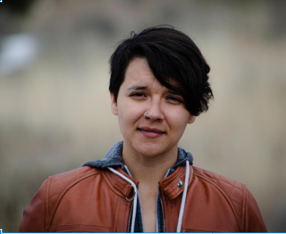 Medium portrait of a Steph smiling and squinting into the camera. With short hair swept across the face. They are wearing a red leather coat over a great hoodie.