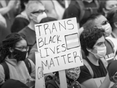 black and white photo of masked protestors. one holds a sign reading "Trans Black Lives Matter."