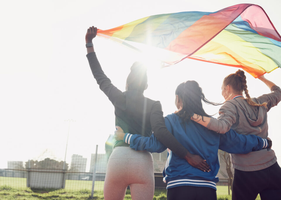 Three people embrace each other while lifting a rainbow flag up in the air.