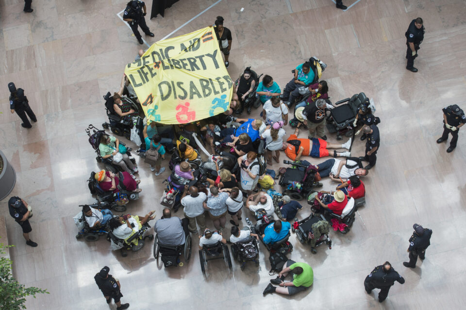 Shot from above, a group of people congregate in a circle in protest turned inward amongst themselves. Some are seated in wheelchairs, or on the ground. They hold up a yellow sign sign that says Medicaid = Life + Liberty 4 Disabled. 