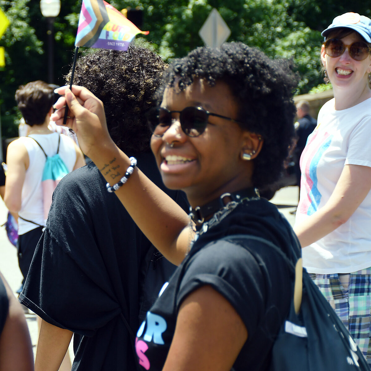 Person holding a trans-inclusive rainbow flag smiles back at the camera.