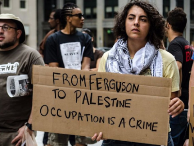 Person at rally holds a sign reading: From Ferguson to Palestine Occupation is a Crime.