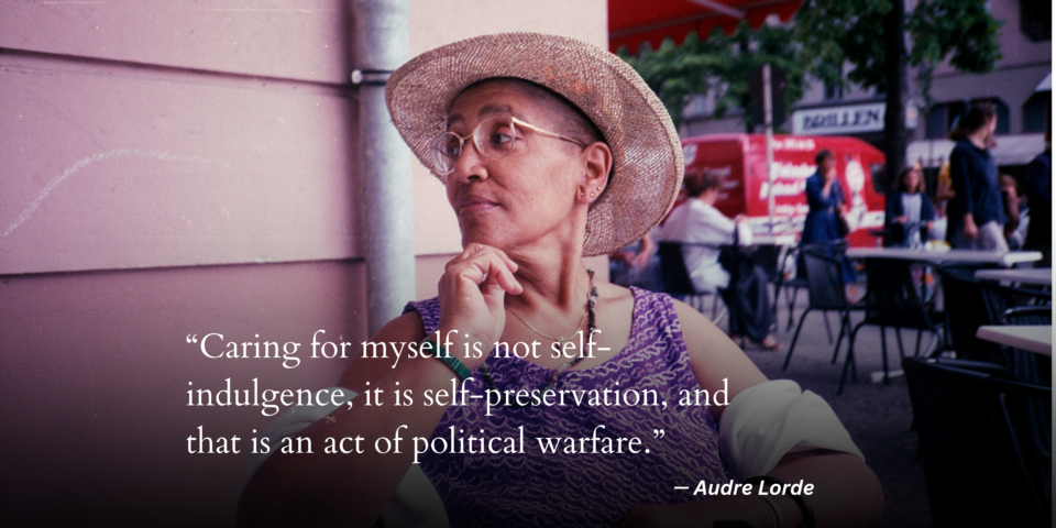 Audre Lorde is seated at the table outdoors with a street view of people walking behind her. She looks knowingly over her shoulder to the left with her hand on her chin. She is wearing a wicker hat, thin gold glasses, a purple top and a string of purple stones around her neck.
