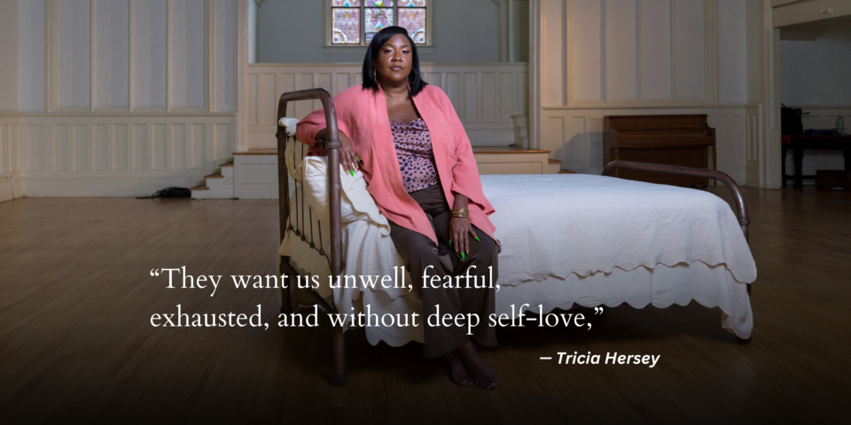 Tricia Hersey sits on top of a bed made in all white linens in the middle of a pewless church with stained glass window in the back; her bare feet are on the floor. She stares confidently into the camera propped on a pillow wearing a pink duster, purple leopard print shirt and brown pants. Her nails are painted bright green and she wears gold bracelets on both hands.