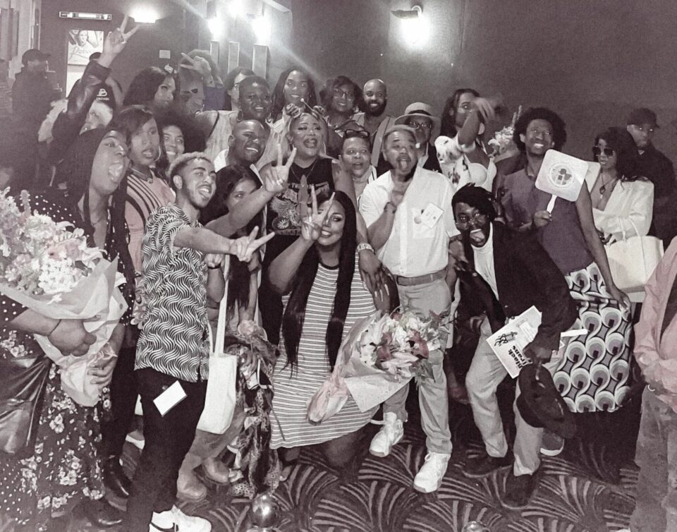 A group photo is captured in black and white of the Lavender Rights Projects team, their community members, and featured comedians at the Black Trans Comedy Showcase, who are each holding a bouquet of followers. A few people are holding up peace signs, and all are smiling