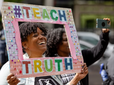 A woman excitedly peers her head into a handmade pink frame with colorful letters around the edges reading "teach truth"