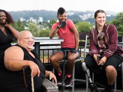 Four disabled people of color with canes and prosthetic legs laugh while chatting. They are on a rooftop deck, in chairs of various height, with greenery and city high-rises in the background.