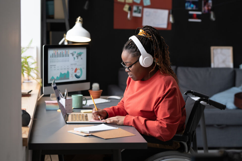 Black woman in braids up in a colorful scarf ponytail, black glasses, and whote headphones, focuses in on her work. She's writing at a computer on a small notepad with a pencil wearing a long-sleeved orange sweatshirt.
