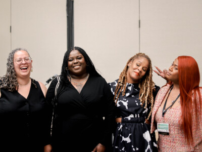 From Left to right: Racial Equity in Inclusion Program Director Alicia Bell, FTG Program Director Dominique Morgan, Shawnda Chapman of Ms. Foundation for Women, and Deshaneir King of Women on the Rise laugh and smile into the camera. Dominique Morgan, with long red hair, wears a pink patterned blazer. Shawnda Chapman’s hair is in locs and is wearing a black and white blouse. Deshaneir King has long dark braids. Alicia Bell also in greying locs celebrate a successful panel.