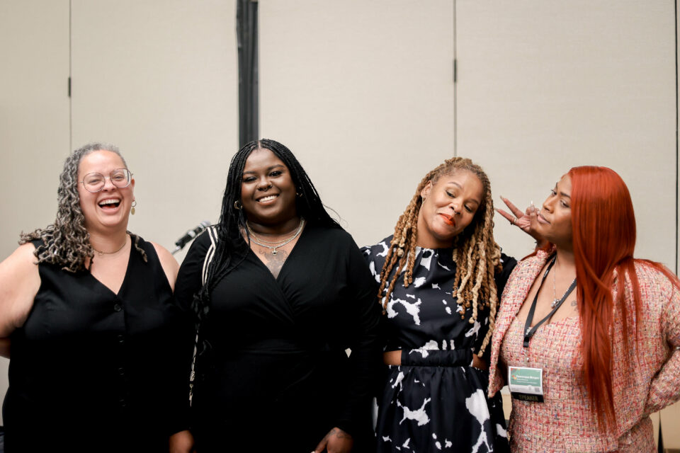 From left to right, Racial Equity in Inclusion Program Director Alicia Bell, Deshaneir King of Women on the Rise, Shawnda Chapman of Ms. Foundation for Women, and FTG Program Director Dominique Morgan, are standed and posed laughing , holding up piece sign, and “smizing” at the camera. Dominique Morgan, with long red hair, wears a pink patterned blazer. Shawnda Chapman’s hair is in locs and is wearing a black and white blouse. Deshaneir King has long dark braids, And Alicia Bell wears a black sleeveless dress.