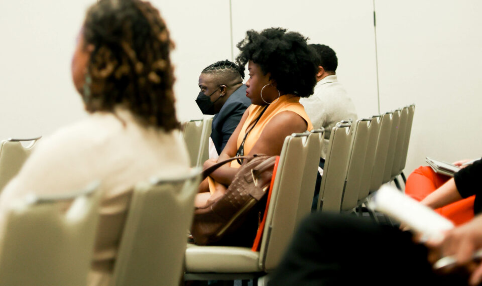 Audience members watch ahead rapt with attention at the presenters outside of the frame. The woman most in focus has a orange shirt and black lanyard and high, black afro hair.