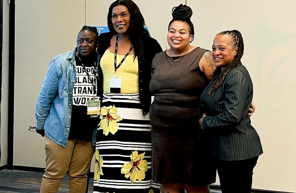 From left to right: TC Caldwell of The Knights & Orchids Society, Zakia McKensey of Nationz Foundation, Nichelle Brunner of ELLC Fund, and Cynthia Renfro of Laughing Gull Foundation posing and smiling together. TC Caldwell is wearing a denim shirt and khaki pants, Zakiya McKenzie is in a black cardigan and yellow floral dress, Nichelle Brunner is in a brown dress, and Cynthia Renfro is in a dark pinstriped suit.