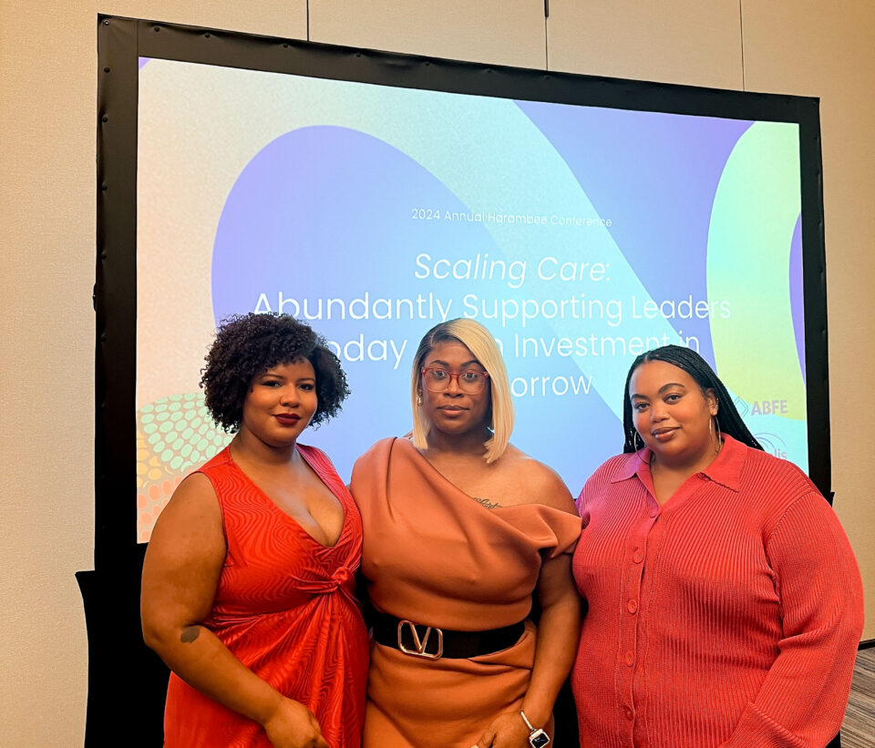 From left to right, Sade Dumas, Dominique Morgan, and Nichelle Brunner, Program Directors of the SJF, FTG, and ELLC Fund, stand in front of a presentation screen at the 2024 Annual Harambee Conference. Sade is wearing a red dress, Dominique is in an off-the-shoulder dress with a black belt, and Nichelle is in a red dress