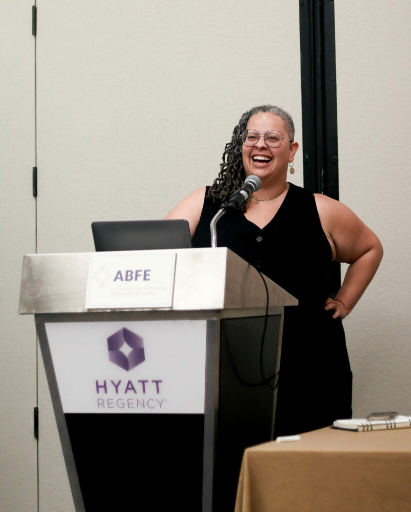 Racial Equity in Inclusion Program Director Alicia Bell, laughs into the microphone at the podium. ABFE and Hyatt Regency written in purple block letters on the front.