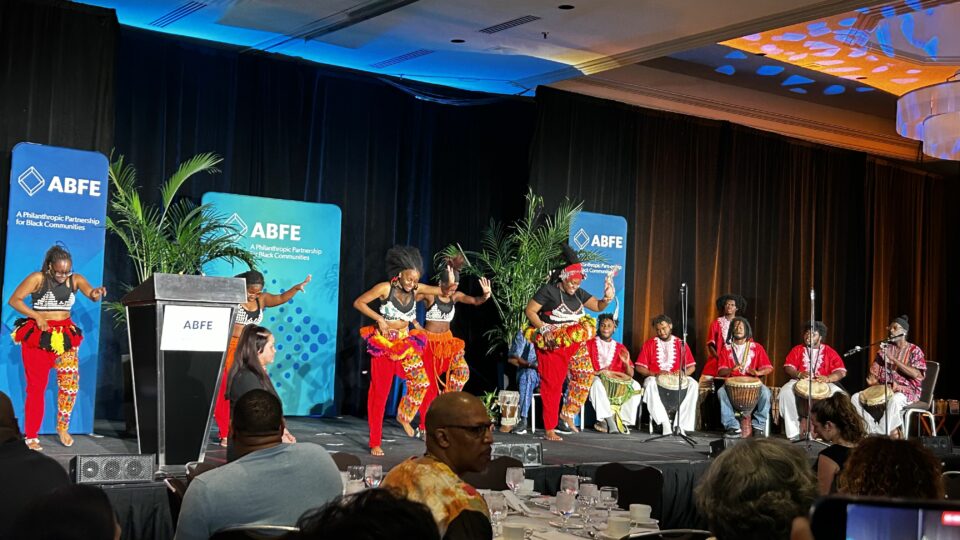 A group of performers are on stage at the ABFE conference. In the foreground, several dancers in brightly colored garments, wearing red pants and vibrant patterns, are dancing on stage. Behind them, musicians dressed in red and white garments are seated playing the drums