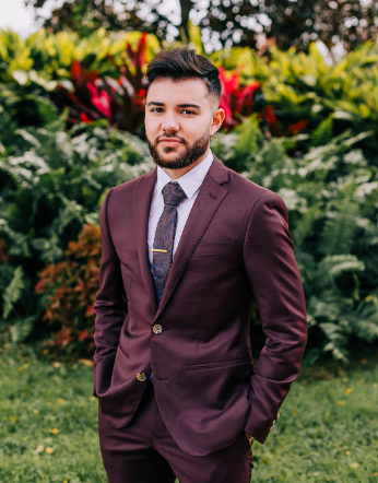 Lucas Gauna stands gazing at the camera in front of a lush backdrop of green red and orange foliage. He is wearing in a maroon-colored suit, pale  lavender shirt and maroon and blue tie with a gold tie pin. His hands are in his pockets; a gold earring in his ear.