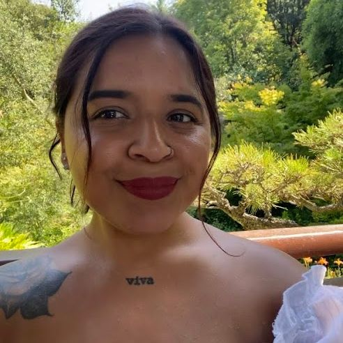 Photo of Arleen Yaz Alonso. She is wearing her hair in an updo, is wearing a nose ring and red lip, and a tattoo that says "viva" (or "alive" in Spanish) is visible on her collar bone.