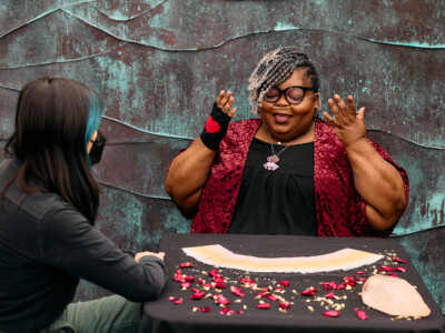 A Black woman gestures while explaining the tarot reading process to a masked genderfluid Asian customer. They are sitting at a table with a tarot deck spread out. The woman wears cat eye glasses, a nasal cannula, and a fingerless glove with a heart in the center on one hand.