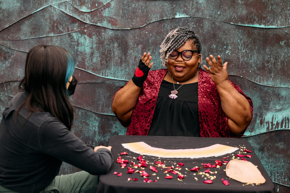 A Black woman gestures while explaining the tarot reading process to a masked genderfluid Asian customer. They are sitting at a table with a tarot deck spread out. The woman wears cat eye glasses, a nasal cannula, and a fingerless glove with a heart in the center on one hand.