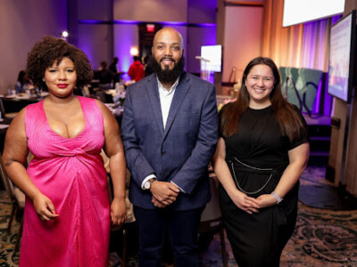 Three individuals are posing together at an event. On the left, a woman is wearing a bright pink dress. In the center, a man with a beard is dressed in a blue suit. On the right, a woman in a black dress is smiling. The background shows a room set up for an event, with tables, chairs, and a screen displaying information. The lighting is warm and the atmosphere appears to be formal and celebratory.