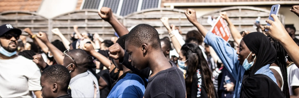 A large group of people, many of whom are wearing face masks, raising their fists in a gesture of solidarity. The crowd includes individuals of various ages and backgrounds, with some holding up phones to capture the moment. The setting appears to be an outdoor public space with buildings in the background/