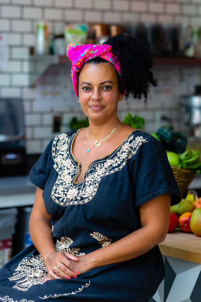 A woman with medium brown skin sits confidently in a well-lit space. She wears a vibrant, pink and multicolored headscarf tied at the front, complementing her curly black hair. Her outfit is a black dress with intricate beige floral embroidery along the neckline and hem. She has a calm, kind expression, with a subtle smile, wearing delicate gold jewelry, including necklaces and rings. Behind her are fresh vegetables and a tiled wall, suggesting a kitchen or cafe environment.