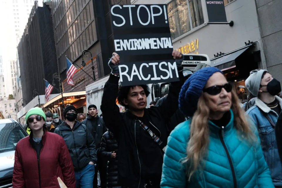 The image shows a diverse group of protesters walking through a city street. A central figure holds up a sign that reads "STOP ENVIRONMENTAL RACISM" in bold, white letters. The individual holding the sign appears serious and determined. People surrounding them, including a person in a blue beanie and a teal jacket, as well as others in casual winter wear, also seem to be participating in the march or protest. In the background, tall buildings and American flags are visible, along with storefront signs, indicating this is an urban setting. Many of the people are wearing masks, likely suggesting a recent event.