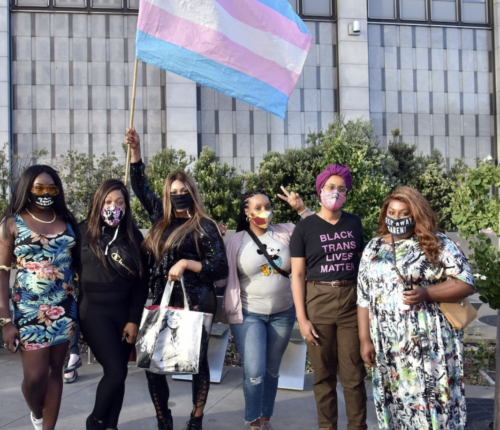 Six people stand together outdoors, wearing face masks and holding a large transgender pride flag. One person’s shirt reads “Black Trans Lives Matter.”