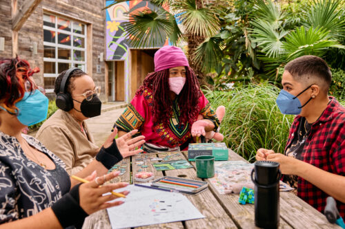 Four people wearing face masks sit together at an outdoor wooden table covered with art supplies, puzzles, and colored pencils, engaged in conversation near a colorful mural and lush greenery.