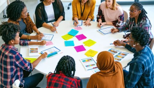 A diverse group of people sit around a circular table engaged in discussion, surrounded by colorful sticky notes, papers, and notebooks during a collaborative workshop or meeting.