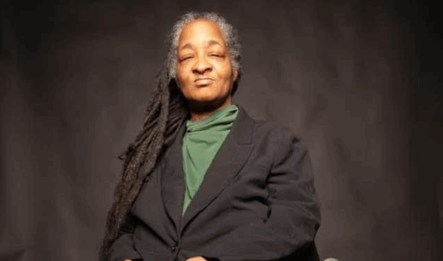 A person with long gray locs and a calm expression sits against a dark backdrop, wearing a green turtleneck and black blazer.