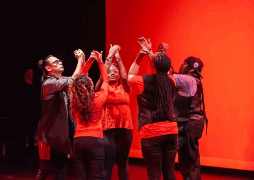 Five performers are standing in a circle on stage, raising their arms and pressing their hands together in a gesture of unity. They're all wearing red and black clothing, with a bold glowing red backdrop behind them.