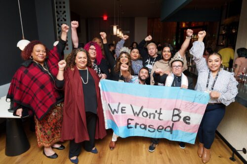 A group of people stand together indoors, raising their fists in solidarity and smiling. They hold a large transgender pride flag with the words “We Won’t Be Erased” written across it. Some individuals wear crowns and lanyards, and the atmosphere appears celebratory and empowering.