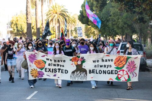 Members of Lavender Phoenix lead a march. They are holding a banner that says “we have always EXISTED” and “we have always BELONGED,” which is decorated with red, orange, and pink flowers and a rooted figure. Behind them, people carry trans flags, Black liberation flags, and protest sign.